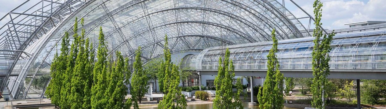 Glasüberdachte Messehalle mit gebogenen Stahlstrukturen, umgeben von Wasserbecken und einer Reihe hoher Bäume.