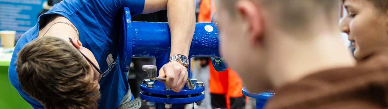 Ein Handwerker zeigt Kinder das befestigen von Rohren.