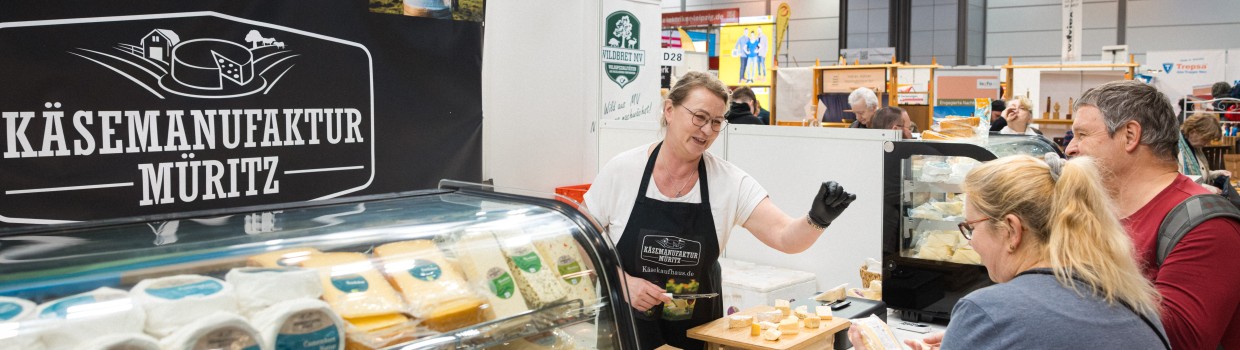 Eine Frau und ein Mann stehen an einem Verkaufsstand für Käse. Die Verkäuferin des Standes berät sie.